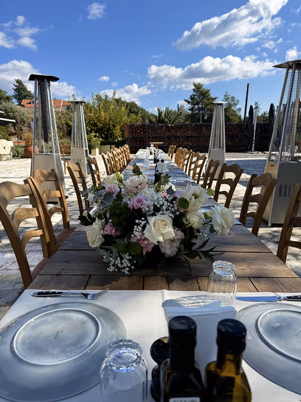 Long outdoor wooden dining table set with plates, glasses, cutlery, and floral centerpiece under blue sky.