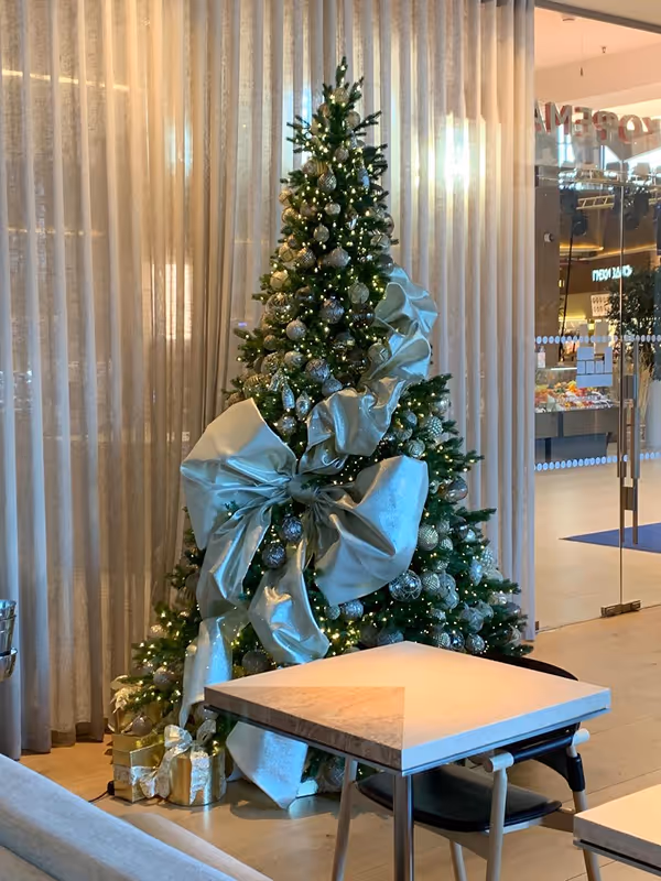 Christmas tree decorated with silver ornaments and a large silver ribbon bow, next to a table and chairs, with wrapped presents underneath.