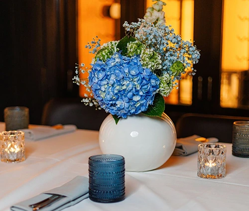 White round vase with blue hydrangea and greenery centerpiece on a white tablecloth surrounded by lit candles and folded napkins.