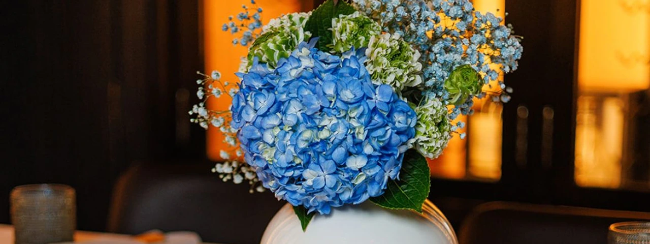 White round vase with blue hydrangea and greenery centerpiece on a white tablecloth surrounded by lit candles and folded napkins.