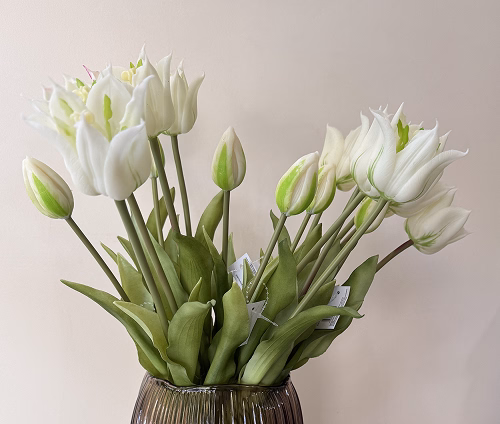A bouquet of white artificial tulips with green stems and leaves in a dark ribbed glass vase