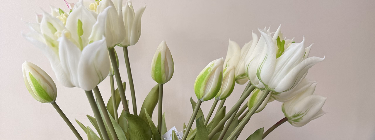 A bouquet of white artificial tulips with green stems and leaves in a dark ribbed glass vase