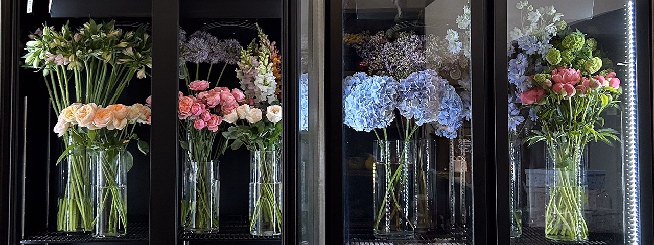 Various fresh flowers in glass vases arranged inside a refrigerated display case.
