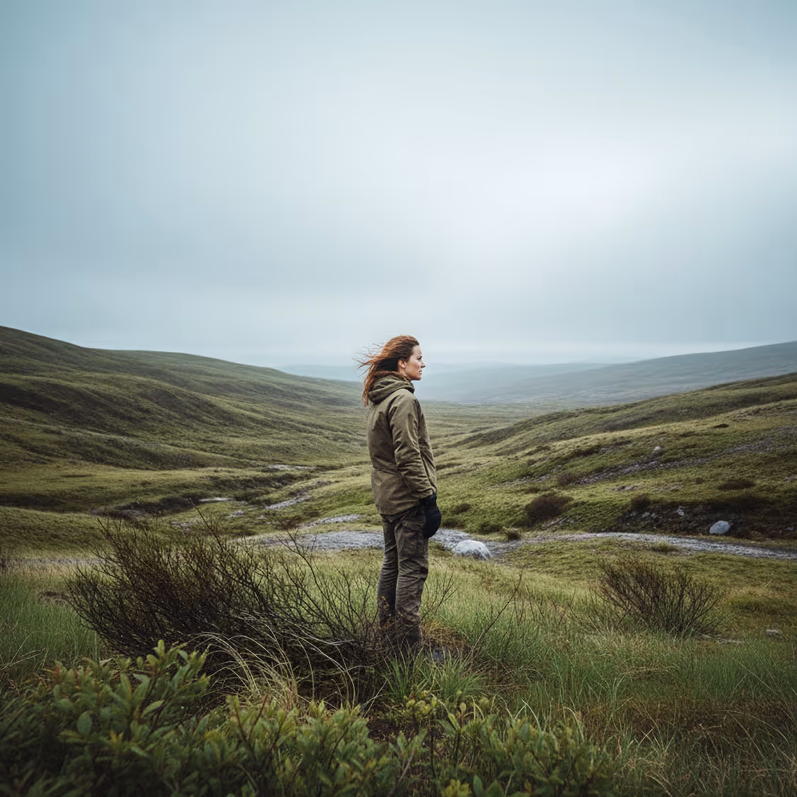 Person in green jacket standing and looking out over expansive green rolling hills under a cloudy sky.