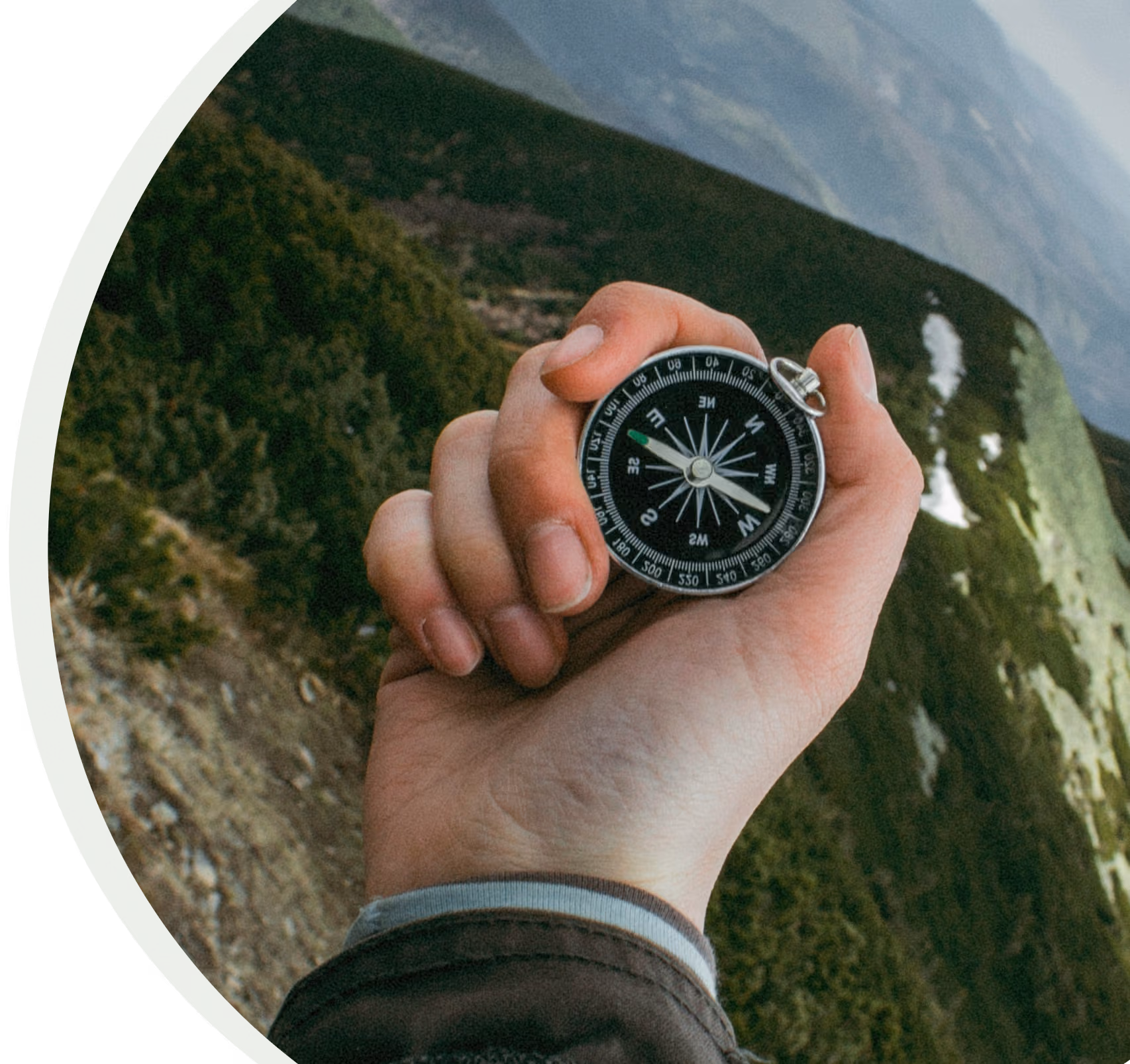 Hand holding a black compass over a mountainous landscape with patches of snow.