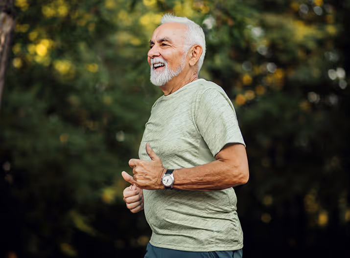 Smiling elderly man jogging outdoors in a green t-shirt with a watch, surrounded by blurred trees.