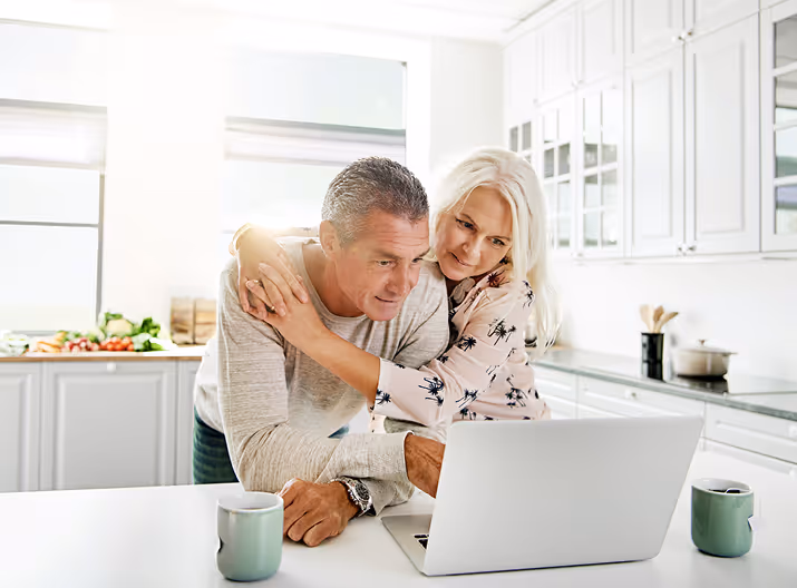 Middle-aged couple standing in a bright kitchen, looking at a laptop with two coffee mugs on the counter.
