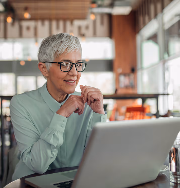 Smiling older woman with short gray hair and glasses working on a laptop in a cozy, well-lit cafe.