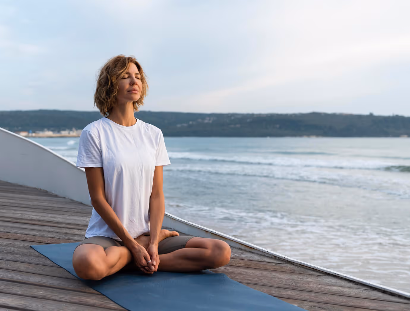 Woman meditating in lotus pose on a yoga mat by the ocean at dawn.
