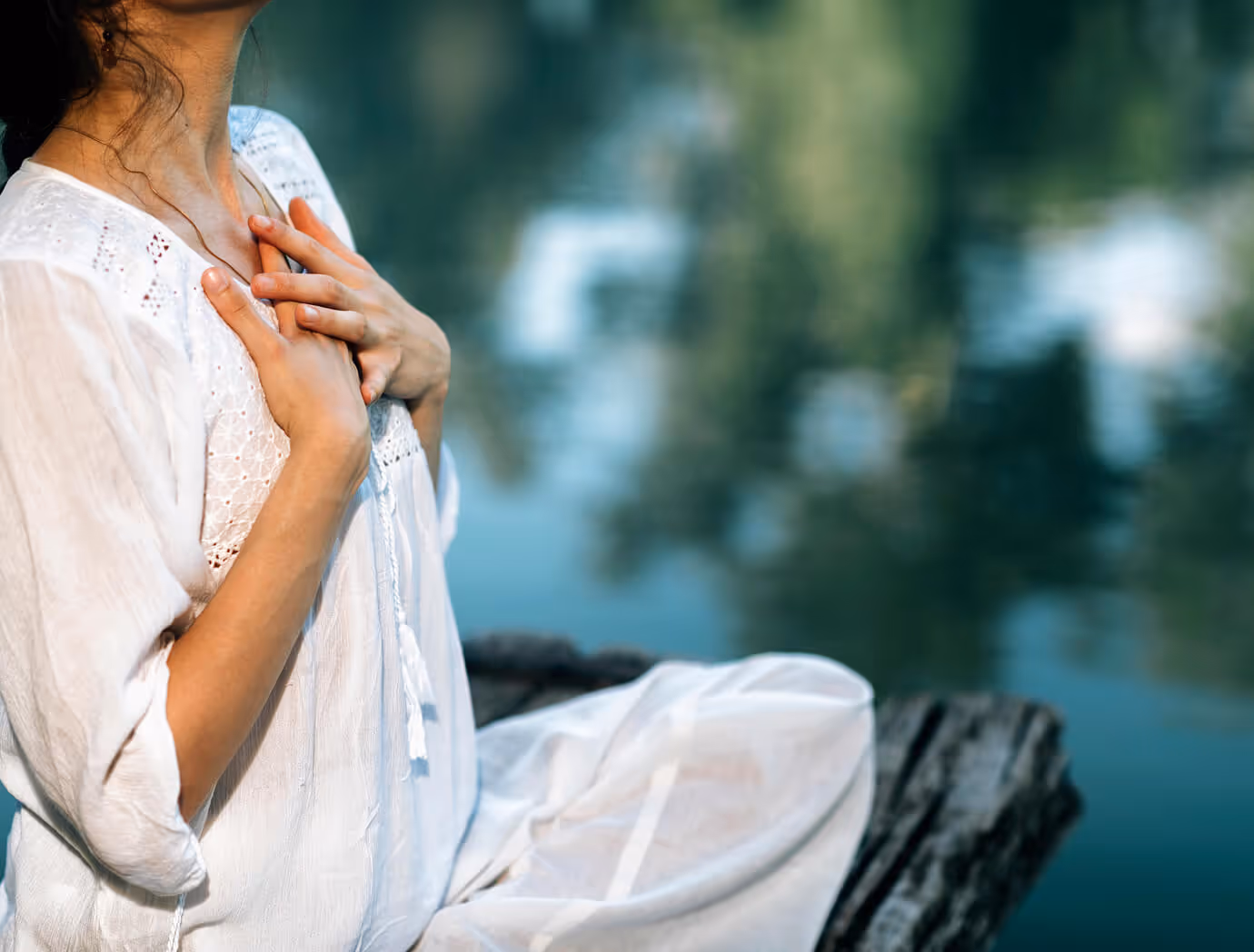 Woman in white dress sitting by water with hands placed on her chest in a peaceful gesture.