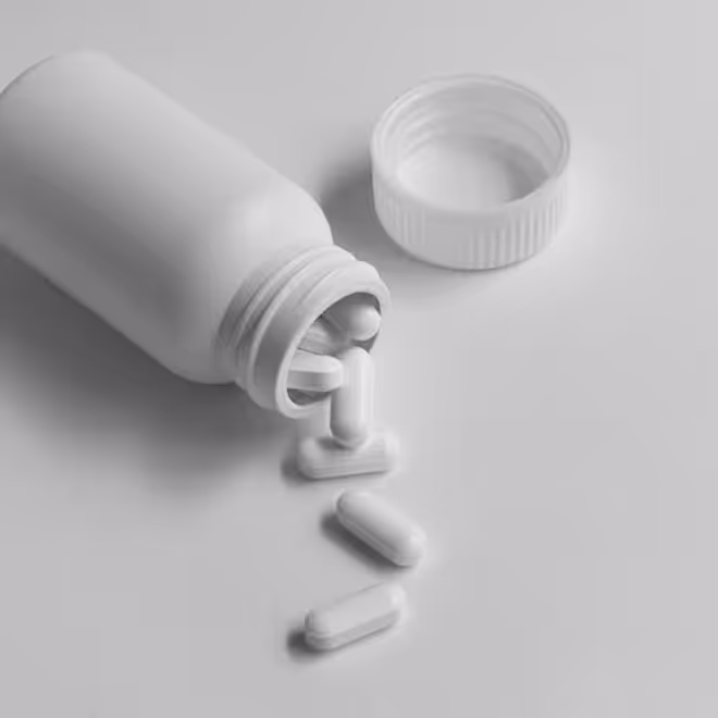 White pill bottle lying on its side with several white capsules spilling out next to the open cap on a white surface.
