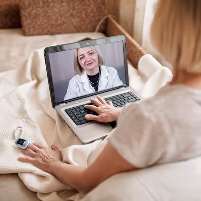 Elderly woman in bed using a laptop for a telehealth video call with a female doctor wearing a white coat on screen.