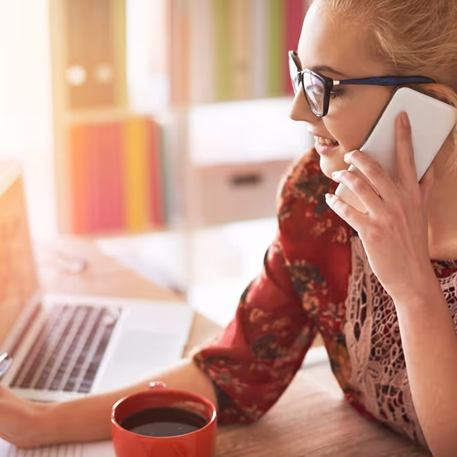 Woman with glasses talking on a smartphone while using a laptop and holding a red coffee cup.