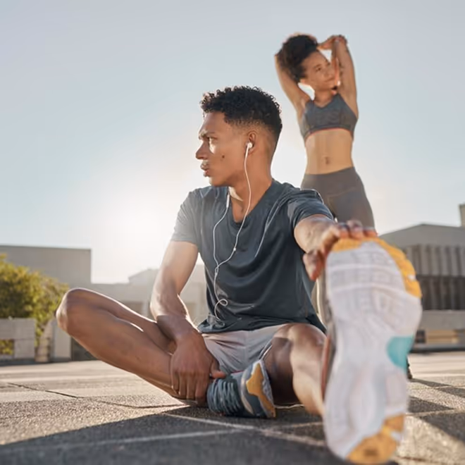 Young man stretching his leg on pavement while a woman in sportswear stretches her arms behind him outdoors.