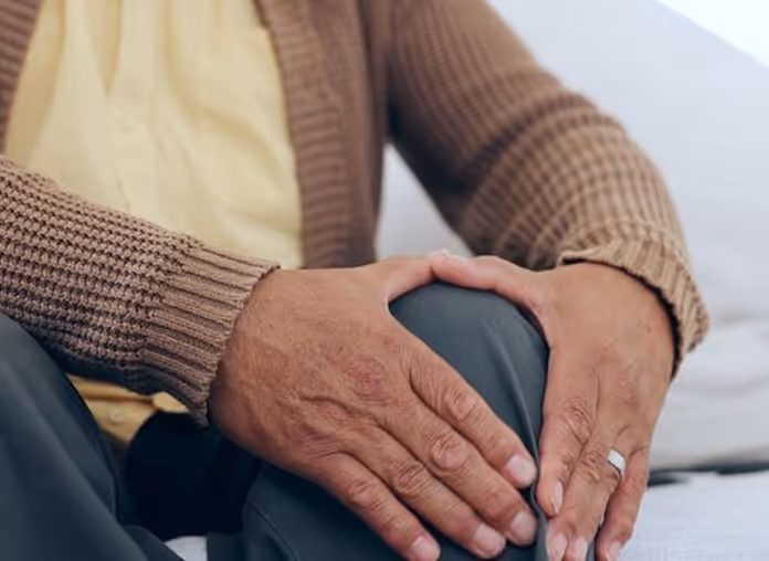 Person wearing a brown sweater and yellow shirt holding their knee with both hands, one with a ring.