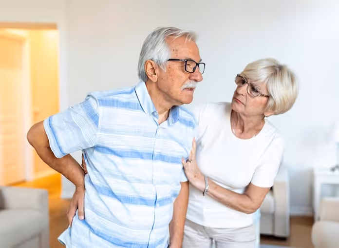 Elderly man in a blue striped shirt holding his lower back in pain while an elderly woman in white looks concerned and supports him.