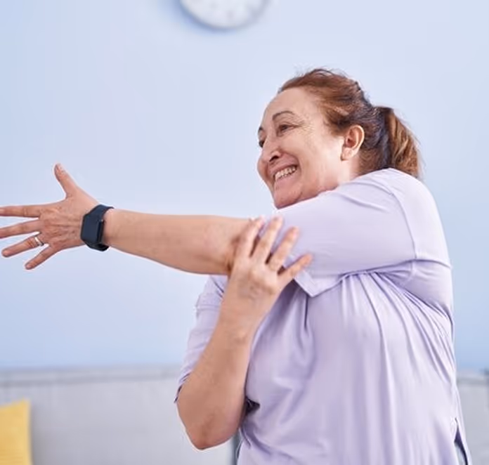 Smiling middle-aged woman in a lavender shirt stretching her arm across her chest in a bright room.
