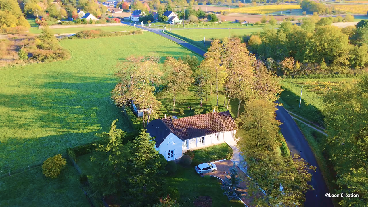 Vue aérienne d'une paisible ferme blanche au toit de tuiles rouges, avec une voiture, des arbres, des champs verdoyants et des routes de campagne sinueuses, baignée dans la lumière chaude de l'automne.