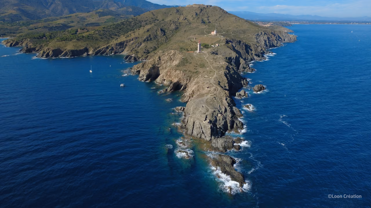 Vue aérienne d'une péninsule côtière au relief accidenté, avec des falaises rocheuses, des collines verdoyantes, un océan bleu, de petites embarcations et un phare, sous une lumière paisible.