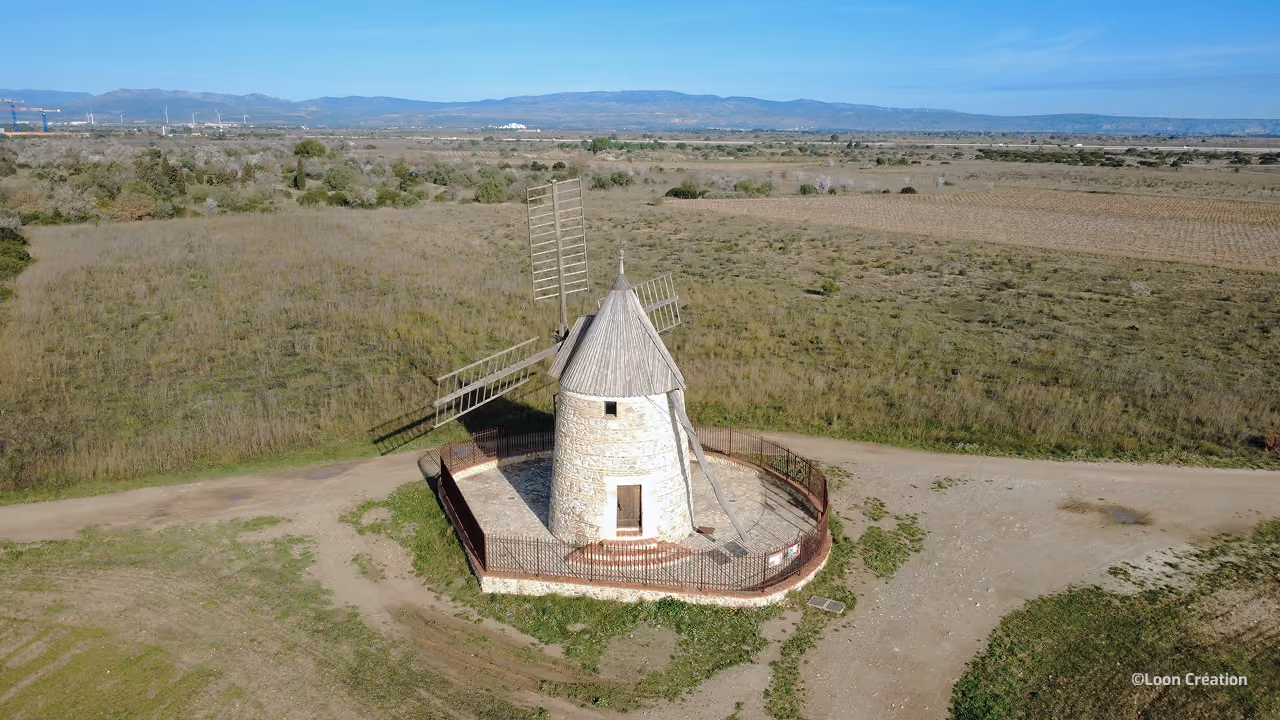 Vue aérienne d'un moulin à vent historique, de Claira, en pierre aux ailes en bois, situé dans un champ rural clôturé et entouré de terres agricoles ainsi que de montagnes bleues au loin.