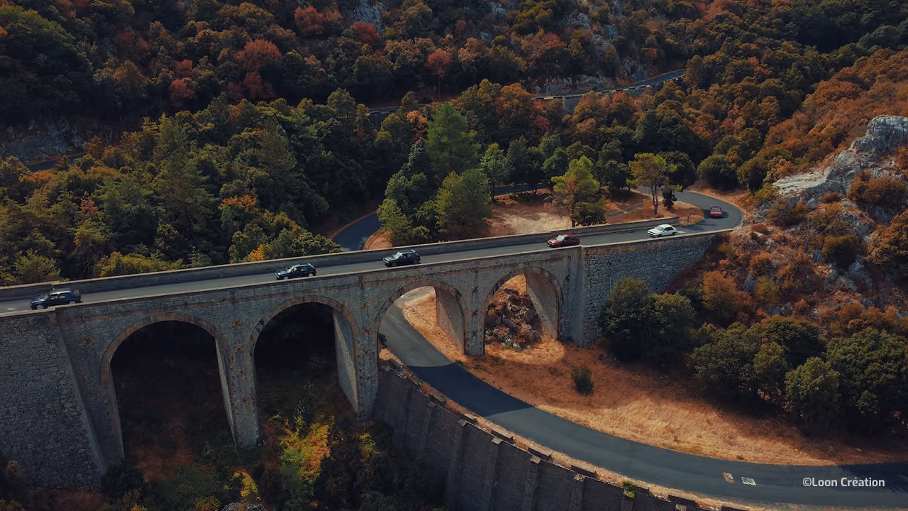 Un viaduc en pierre serpente à travers une forêt aux couleurs automnales, surplombant une route sinueuse ; la vue aérienne dégage une atmosphère paisible et pittoresque.