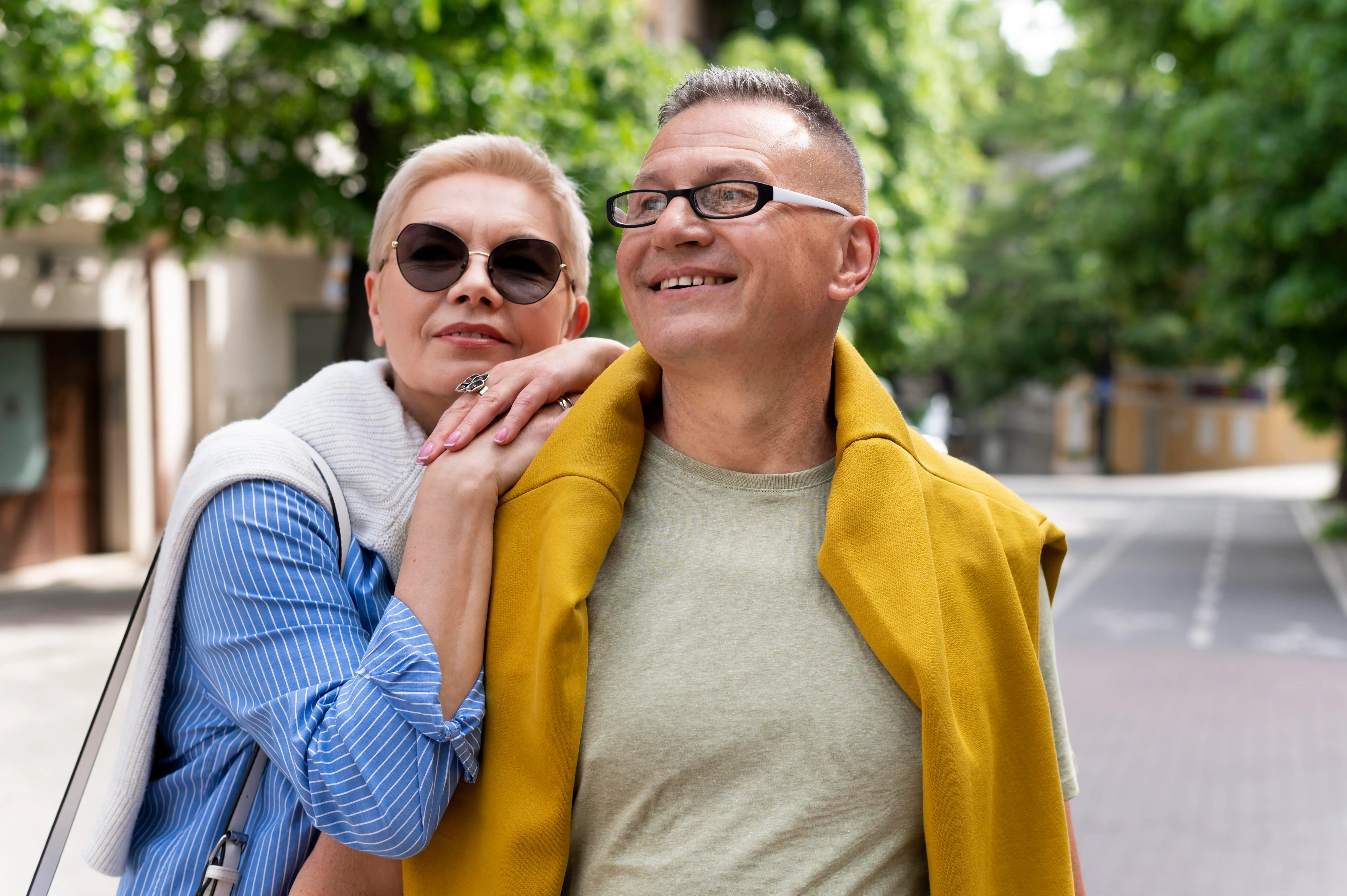 Two adults wearing glasses posing together outdoors on a tree-lined street