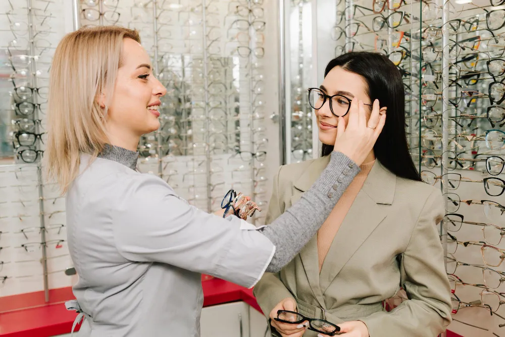 Two women smile at each other in an eyeglasses store, holding glasses displays.