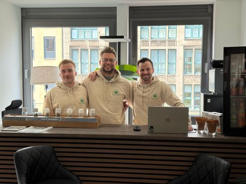 Drei Männer in beige Hoodies mit Cannabislogo stehen hinter einem Tresen mit Probiergläsern und Laptop.