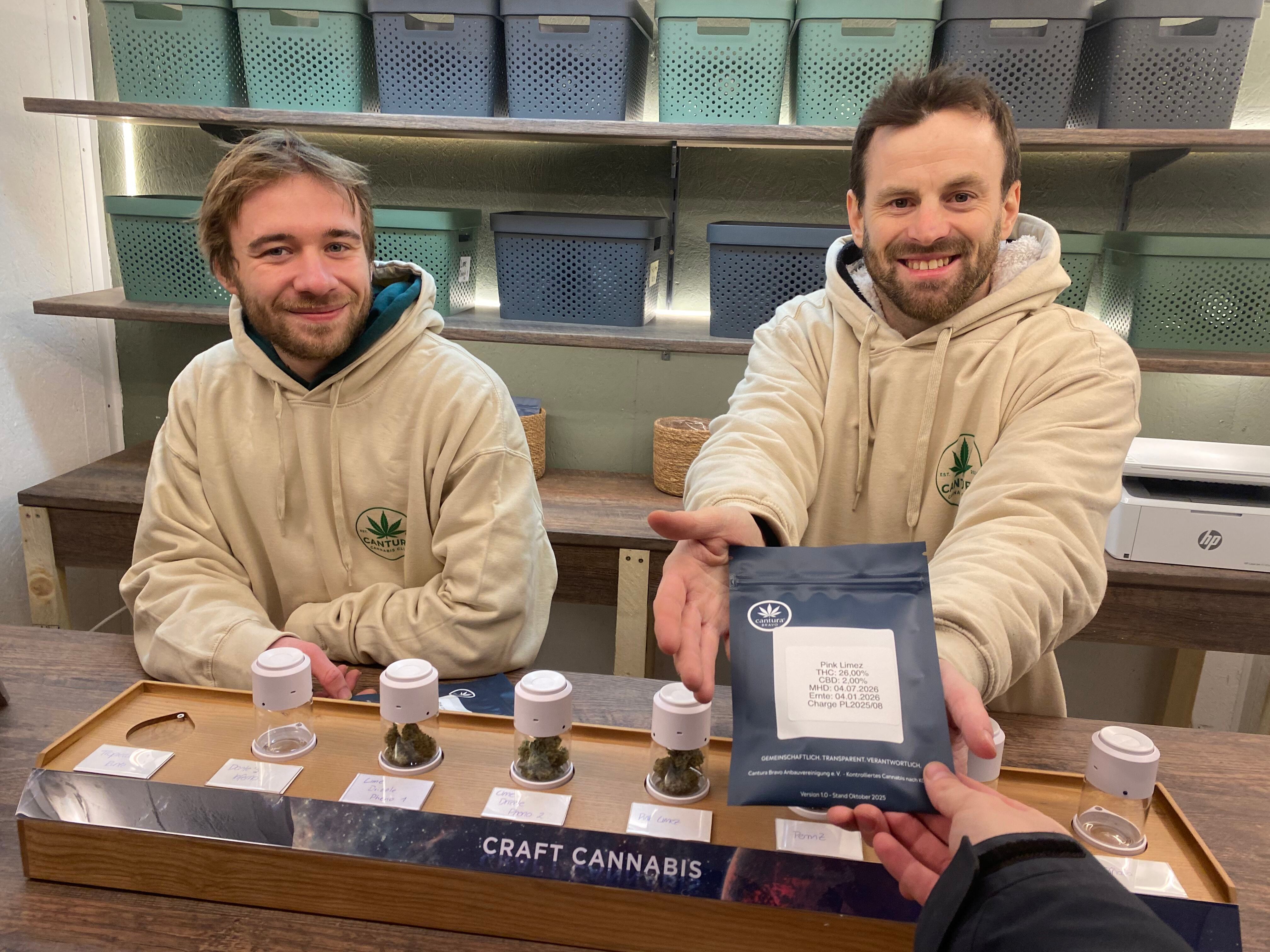 Zwei Männer in beige Kapuzenpullovern präsentieren Cannabissorten und eine Packung Craft Cannabis.