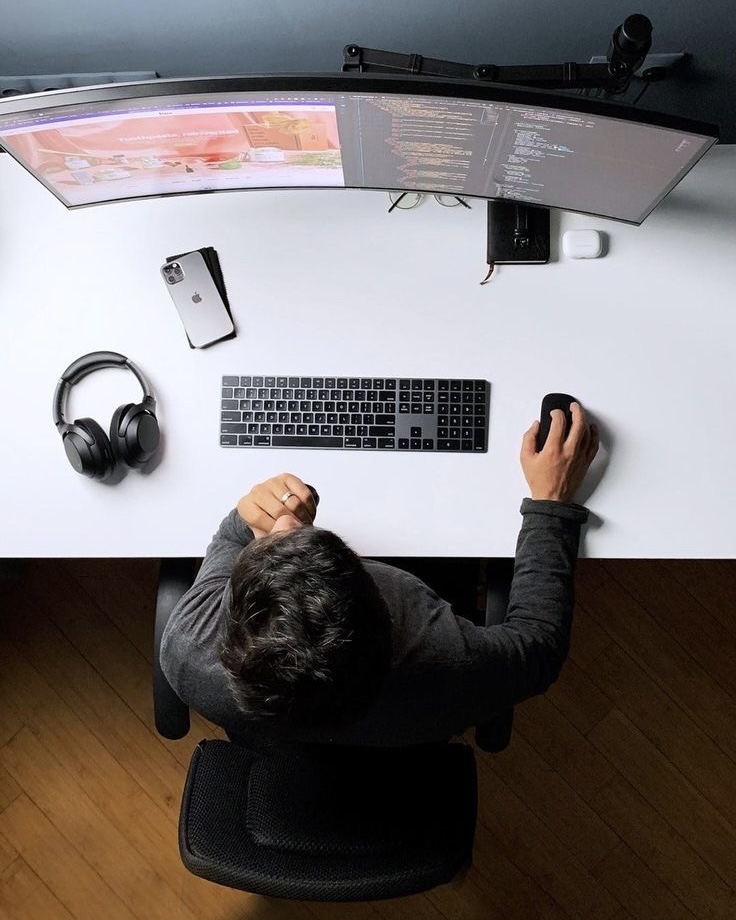 Personne vue de dessus travaillant sur un bureau blanc avec clavier, souris, casque, smartphone et deux écrans affichant du code et un site web.