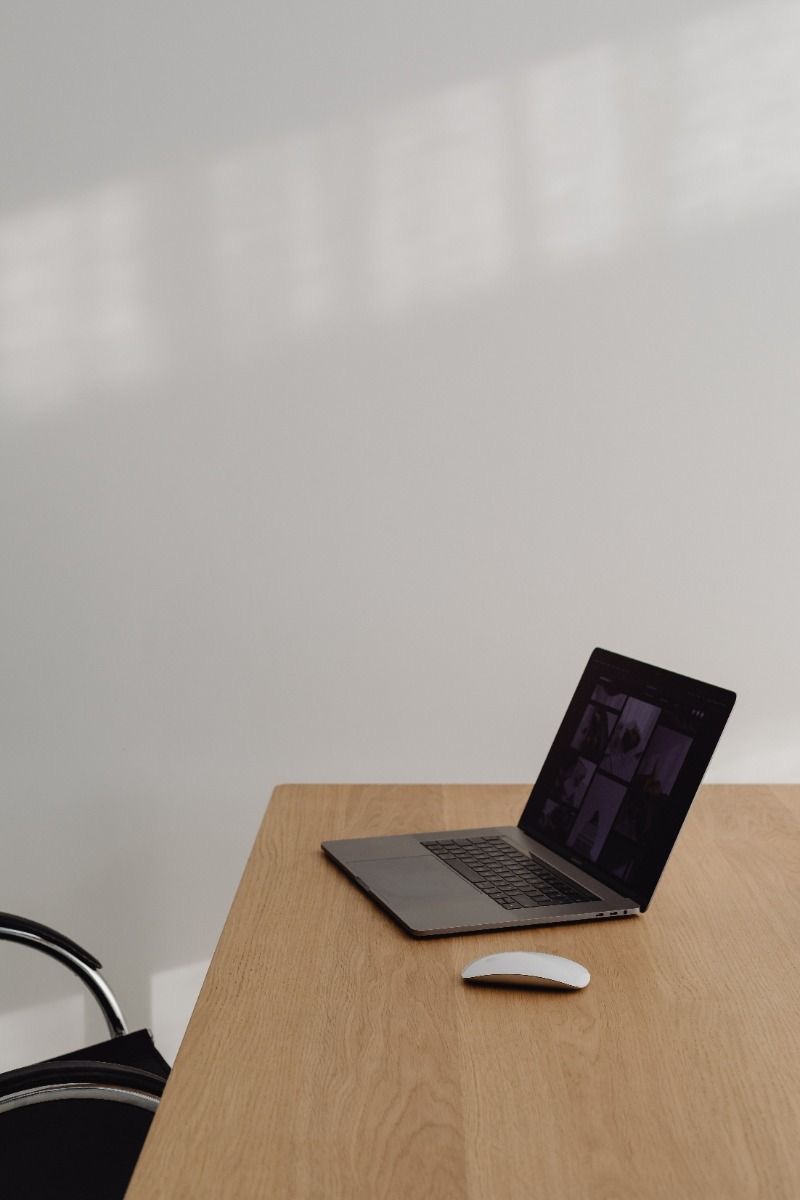 Ordinateur portable ouvert et souris blanche sur une table en bois, chaise noire partiellement visible, fond mur blanc avec ombres.