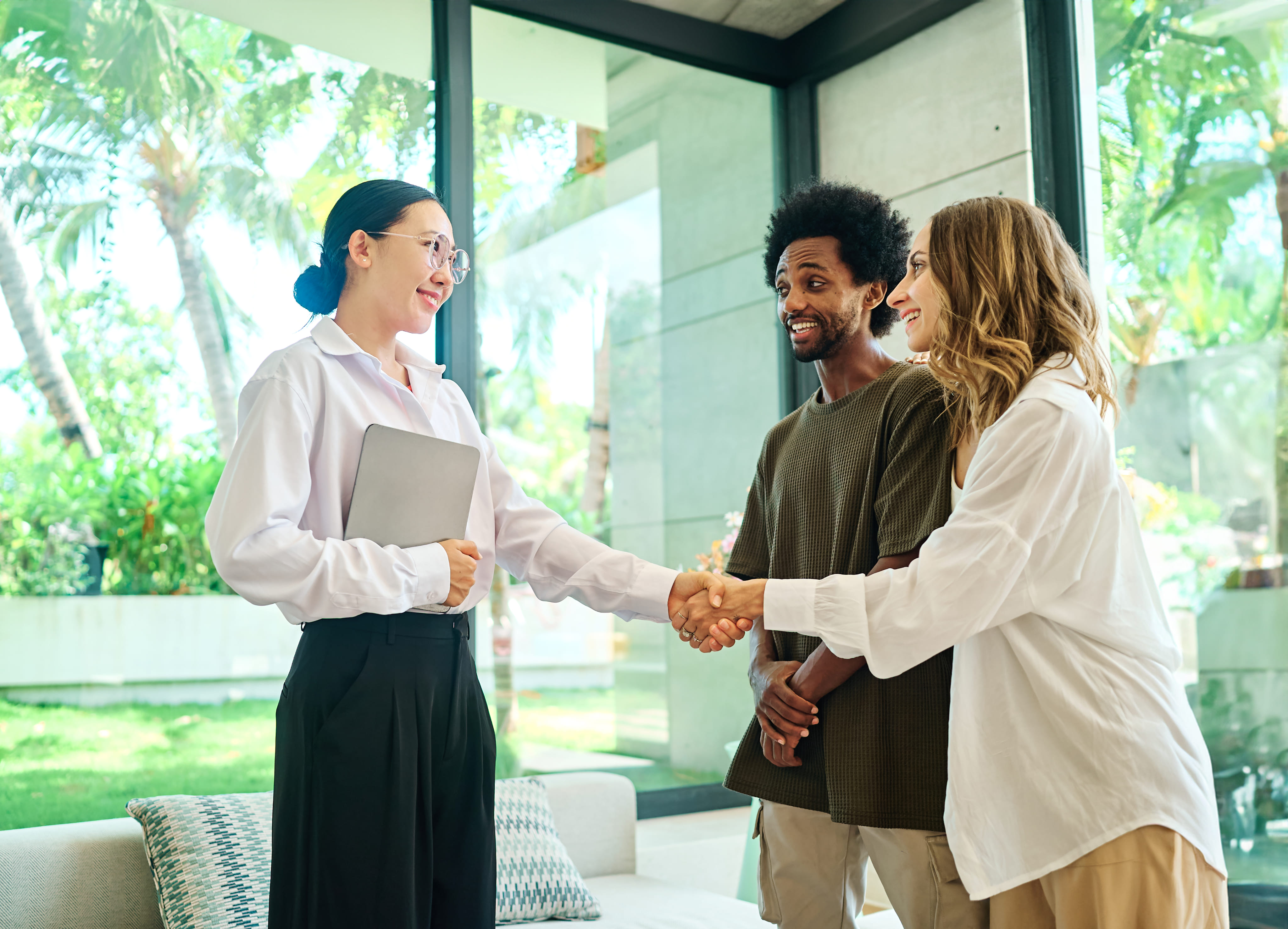 A professional woman holding a digital tablet shakes hands with a smiling couple inside a modern, brightly lit home.