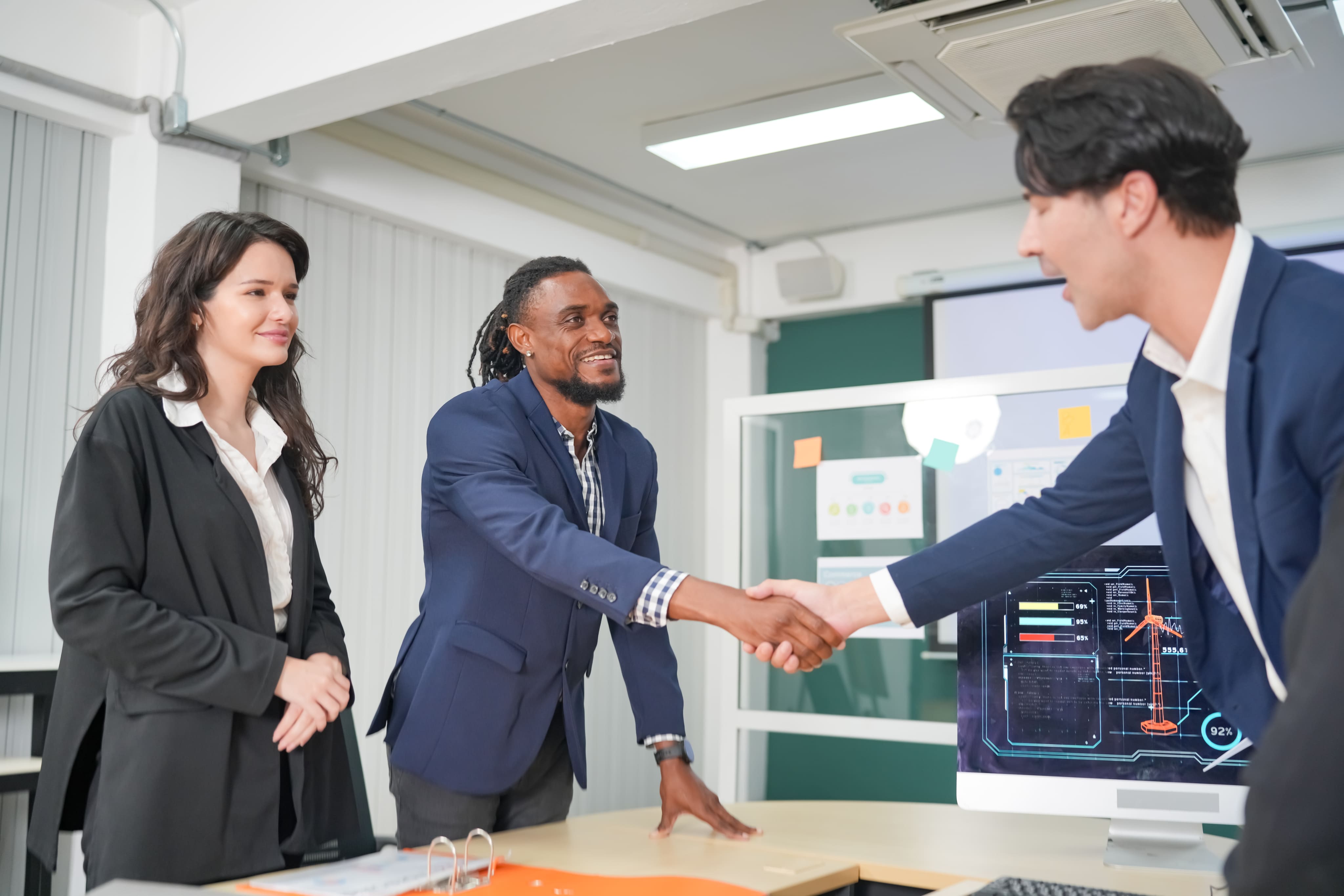 Two businessmen shaking hands across a table during a meeting while a female colleague looks on.