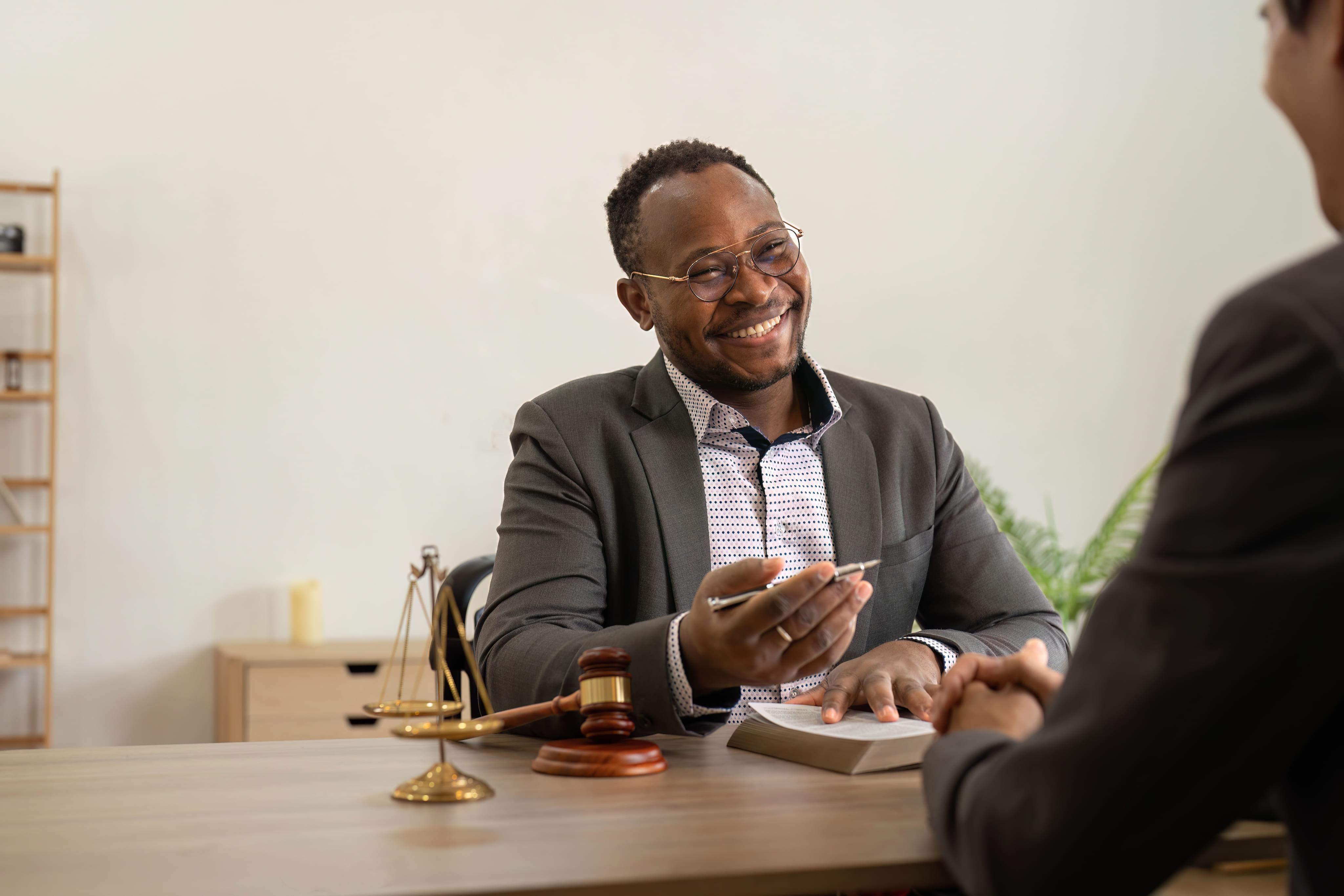 A smiling male attorney wearing glasses sits at his desk during a supportive consultation with a client.