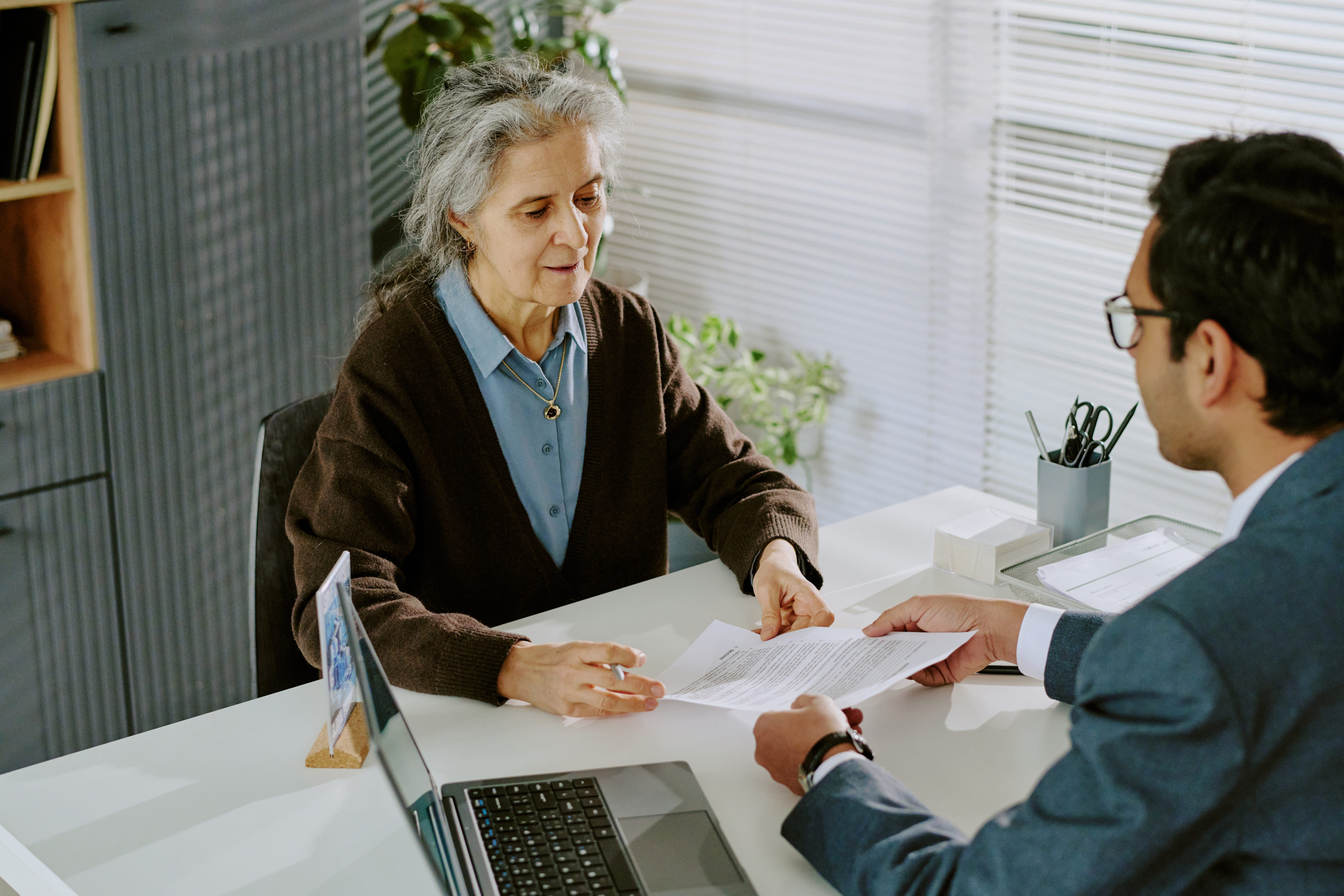 An elderly woman with grey hair sitting at a bright desk reviewing a document with a male legal advisor.