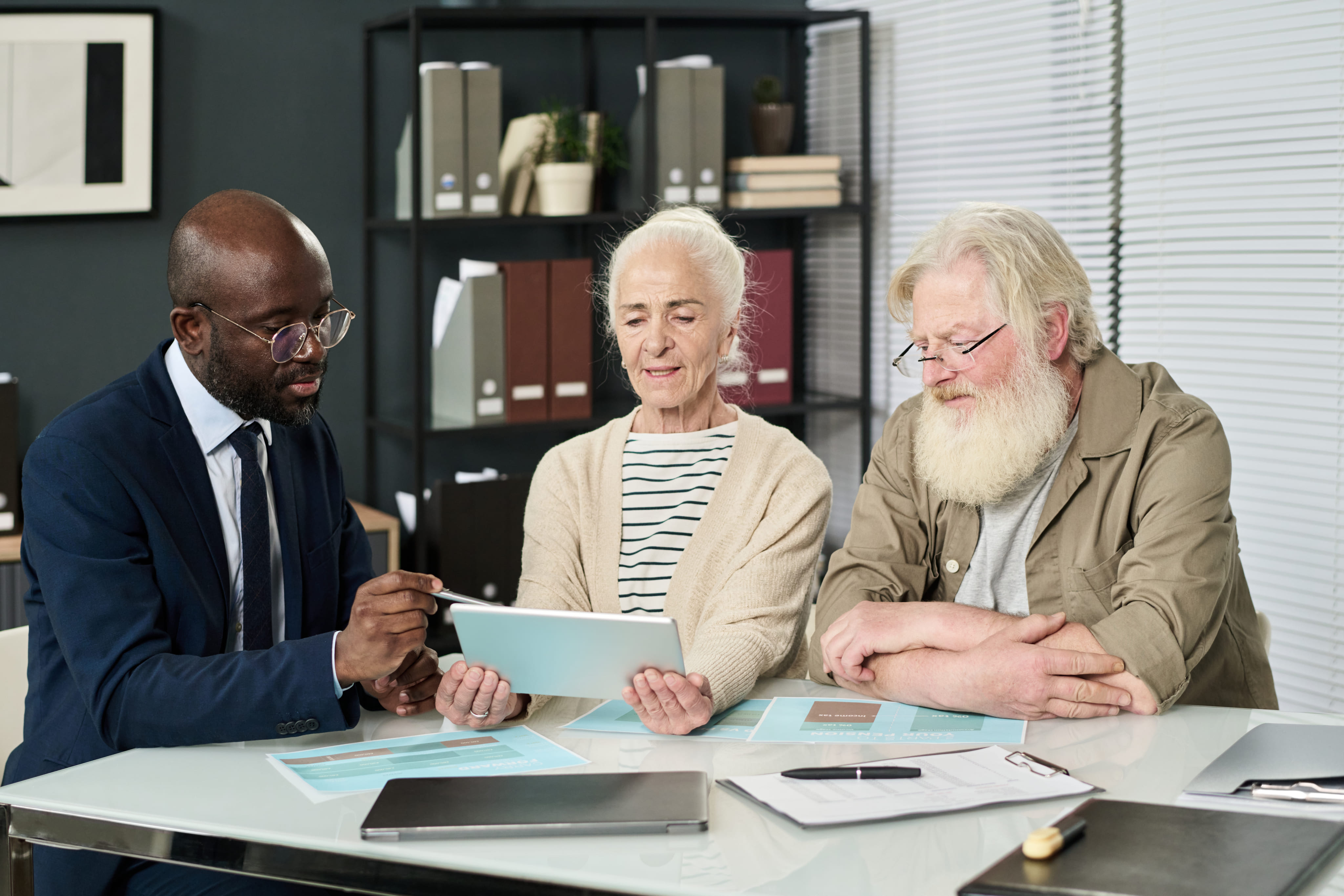 A male attorney in a suit reviewing information on a tablet with an elderly couple in an office.