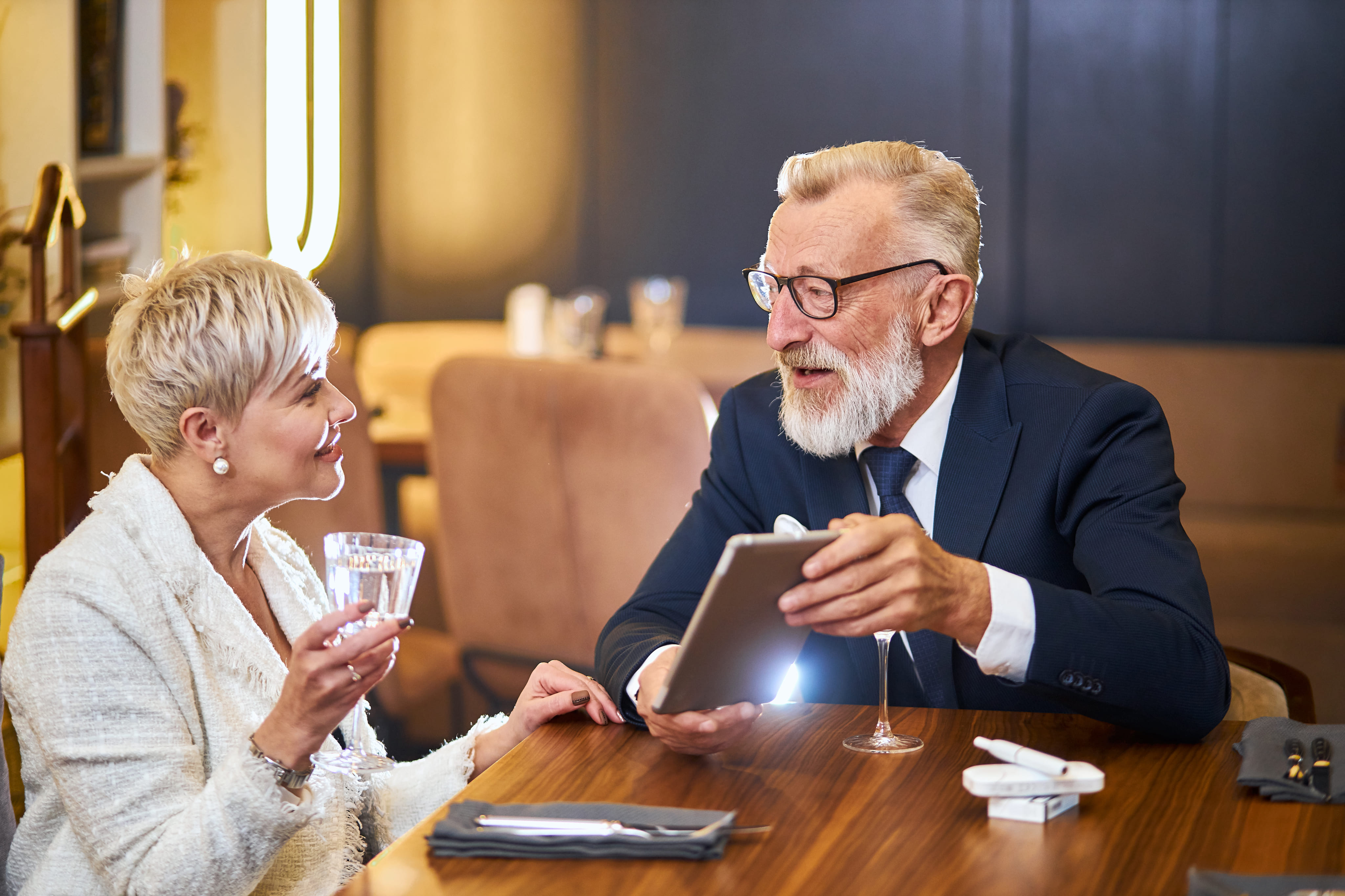 An older couple smiling and looking at a digital tablet together while sitting at a table.