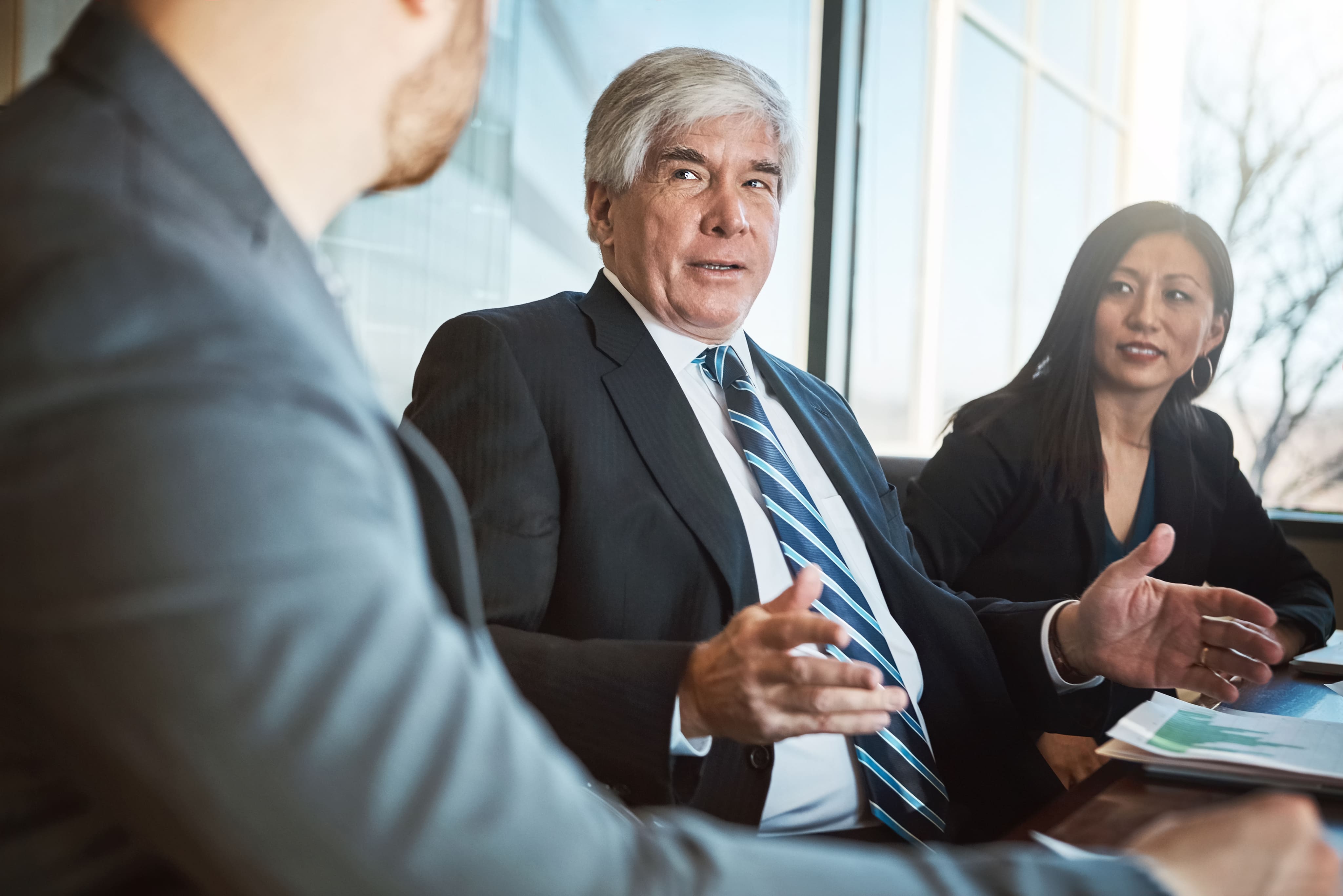 A senior corporate professional with grey hair gesturing during a business meeting with colleagues around a conference table.