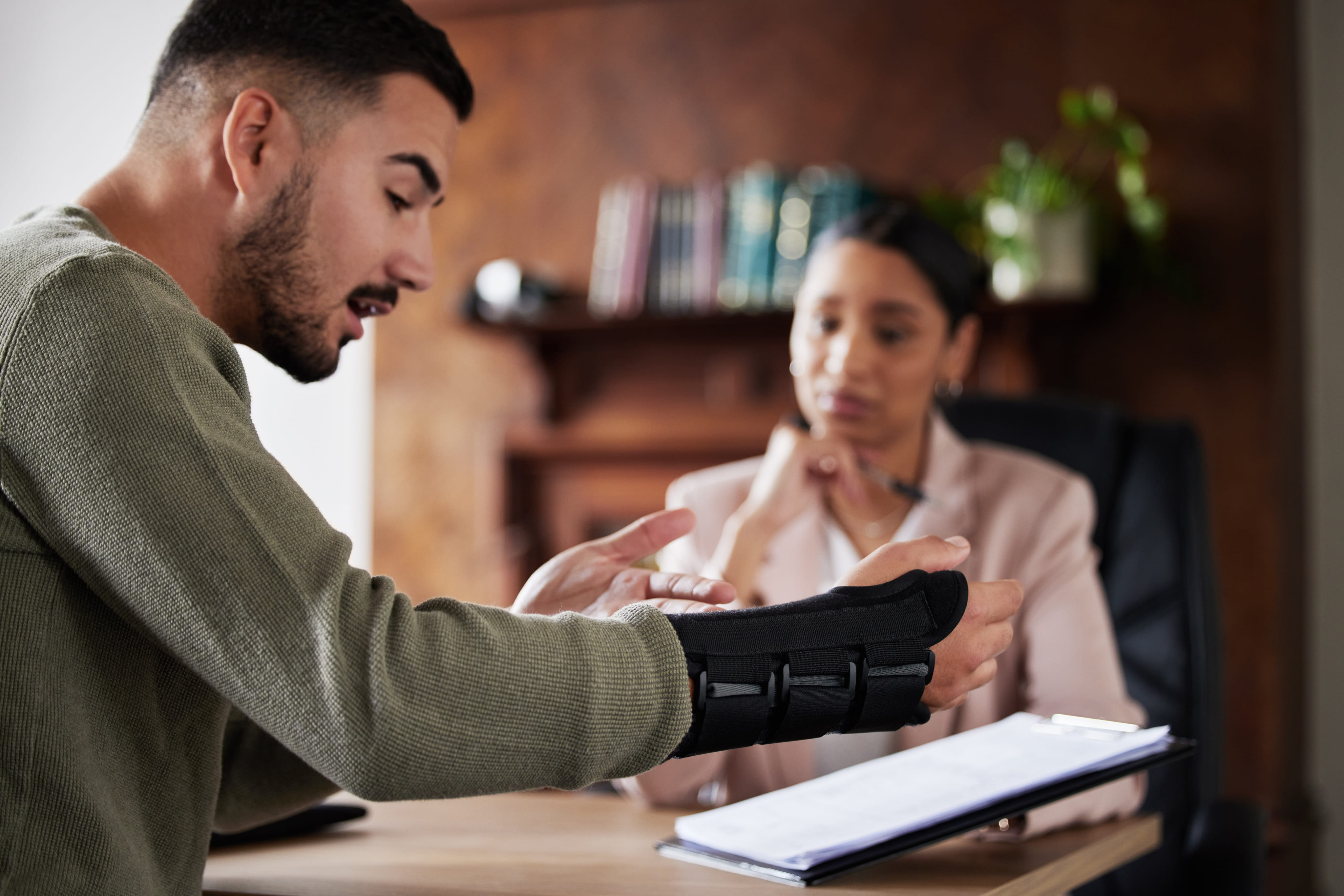 An injured man wearing a black wrist brace discussing his case with a female professional at a desk.