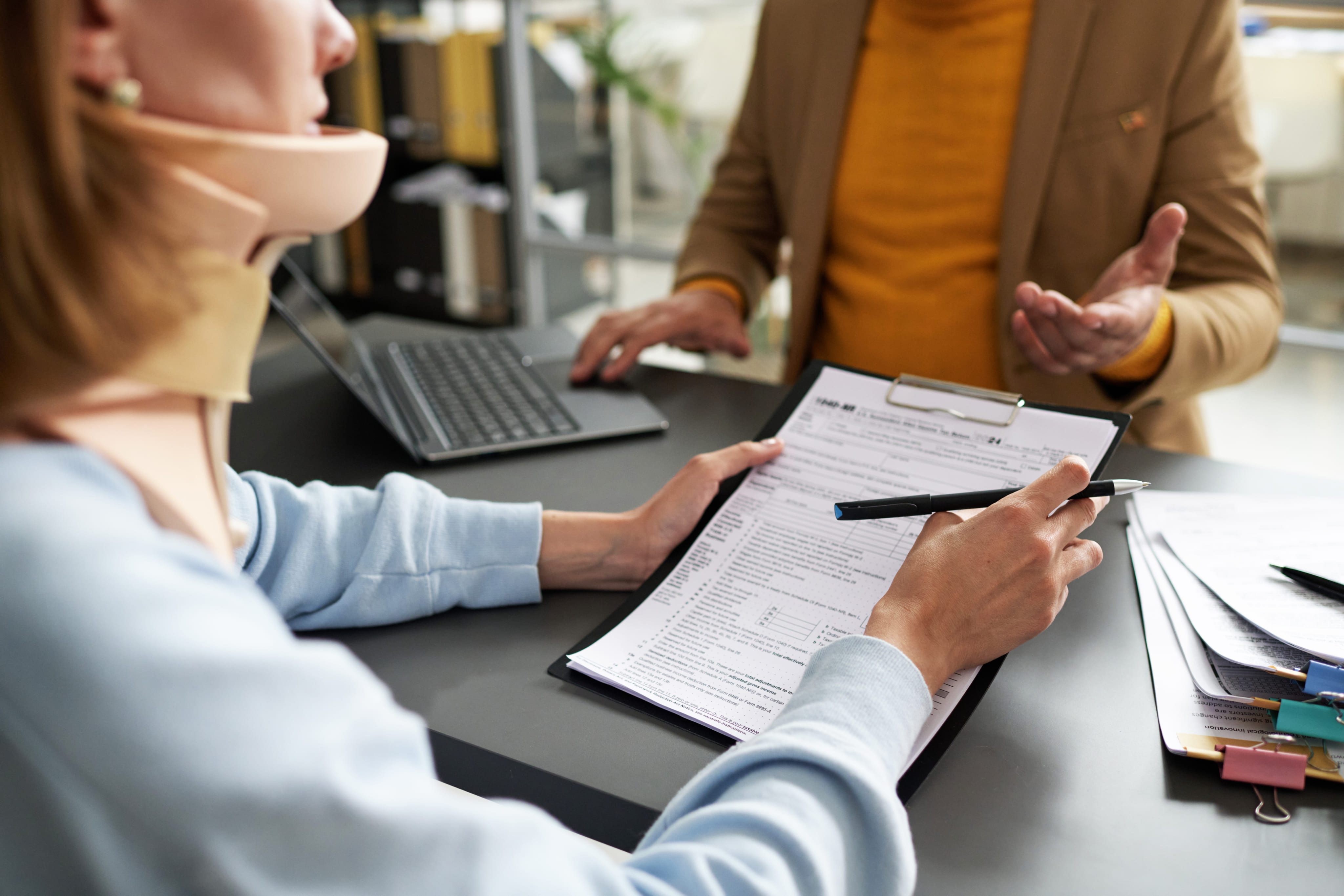 A client wearing a cervical neck collar reviewing legal documents with an attorney at a desk.