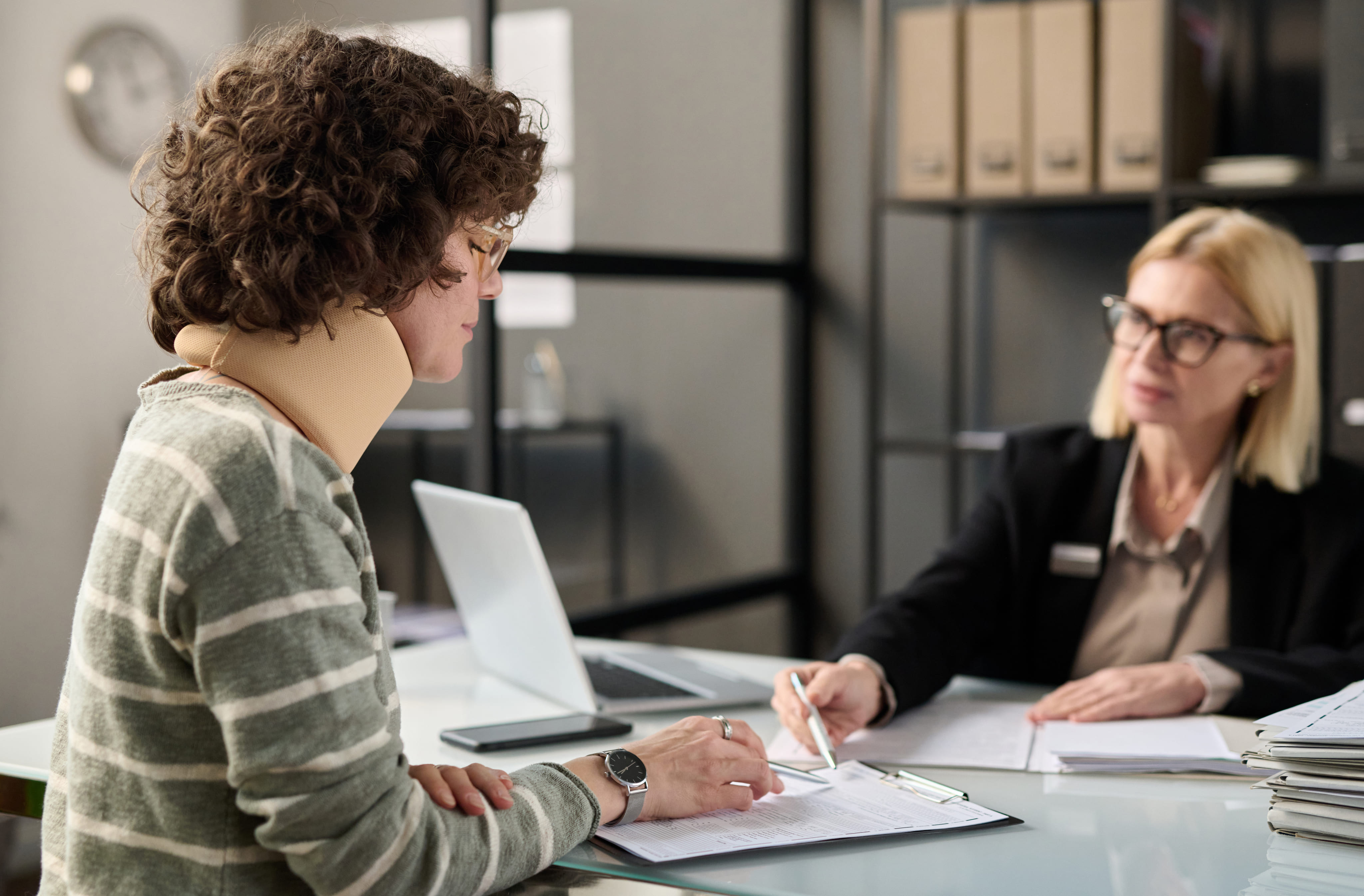 A client wearing a tan neck brace consulting with a blonde female legal professional in an office.