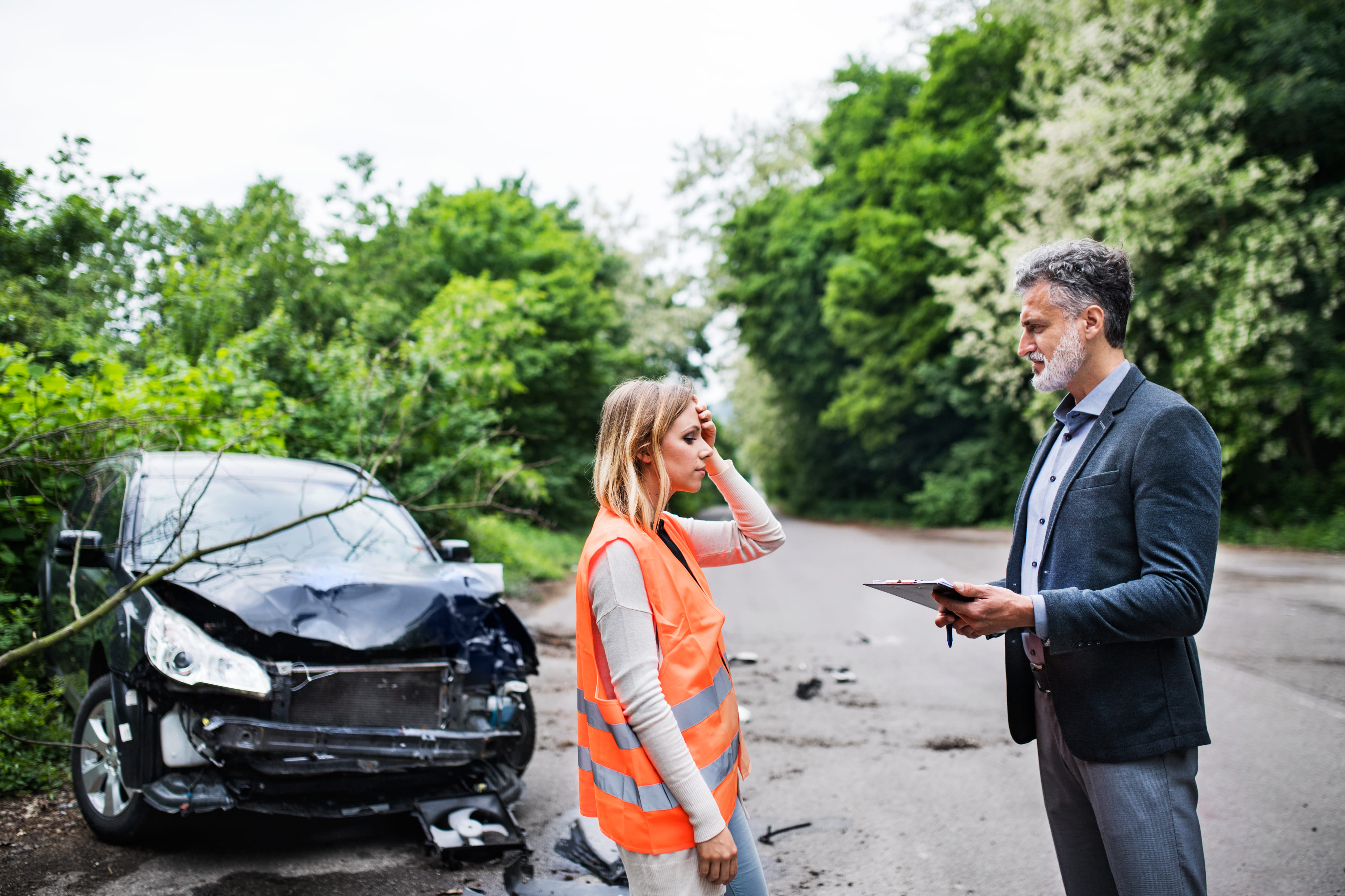 A woman wearing an emergency safety vest talking to a professional next to a heavily damaged car on the side of a road.