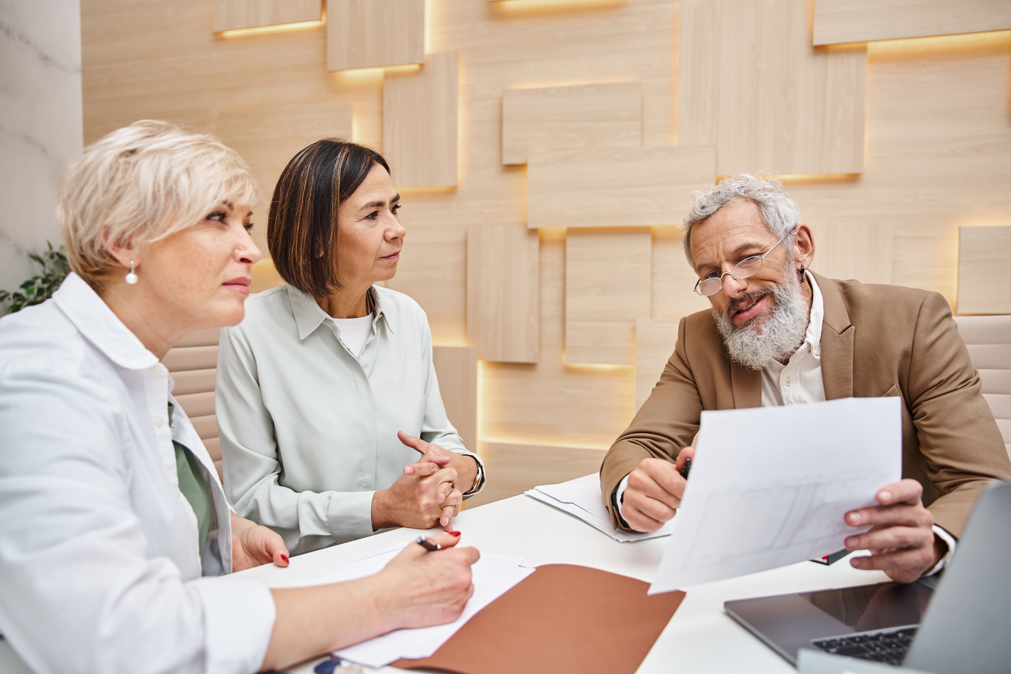 A senior male advisor reviewing estate planning documents with two women around a wooden table.