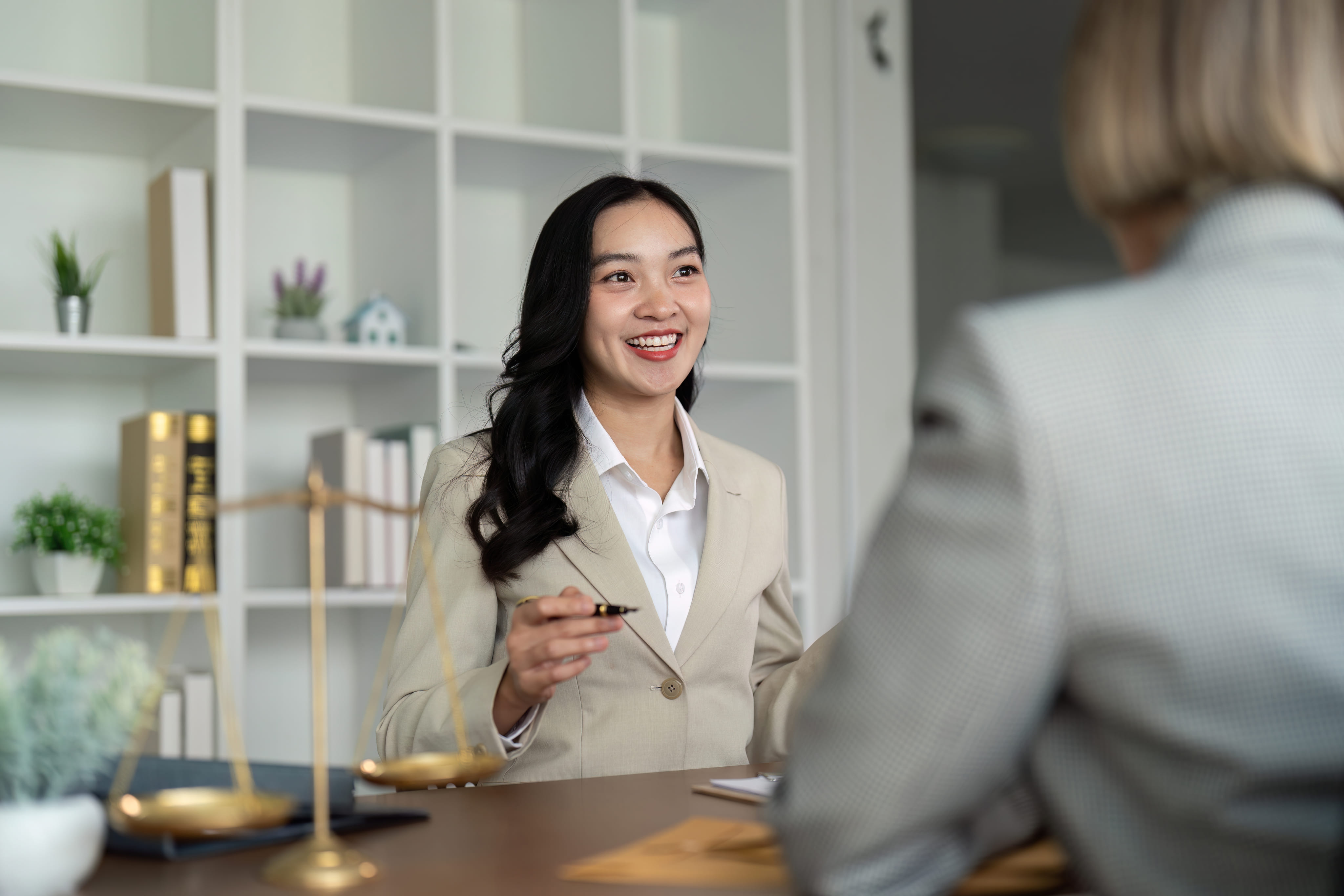 A smiling female legal professional in a light blazer speaking with a client at a desk featuring a scales of justice ornament.