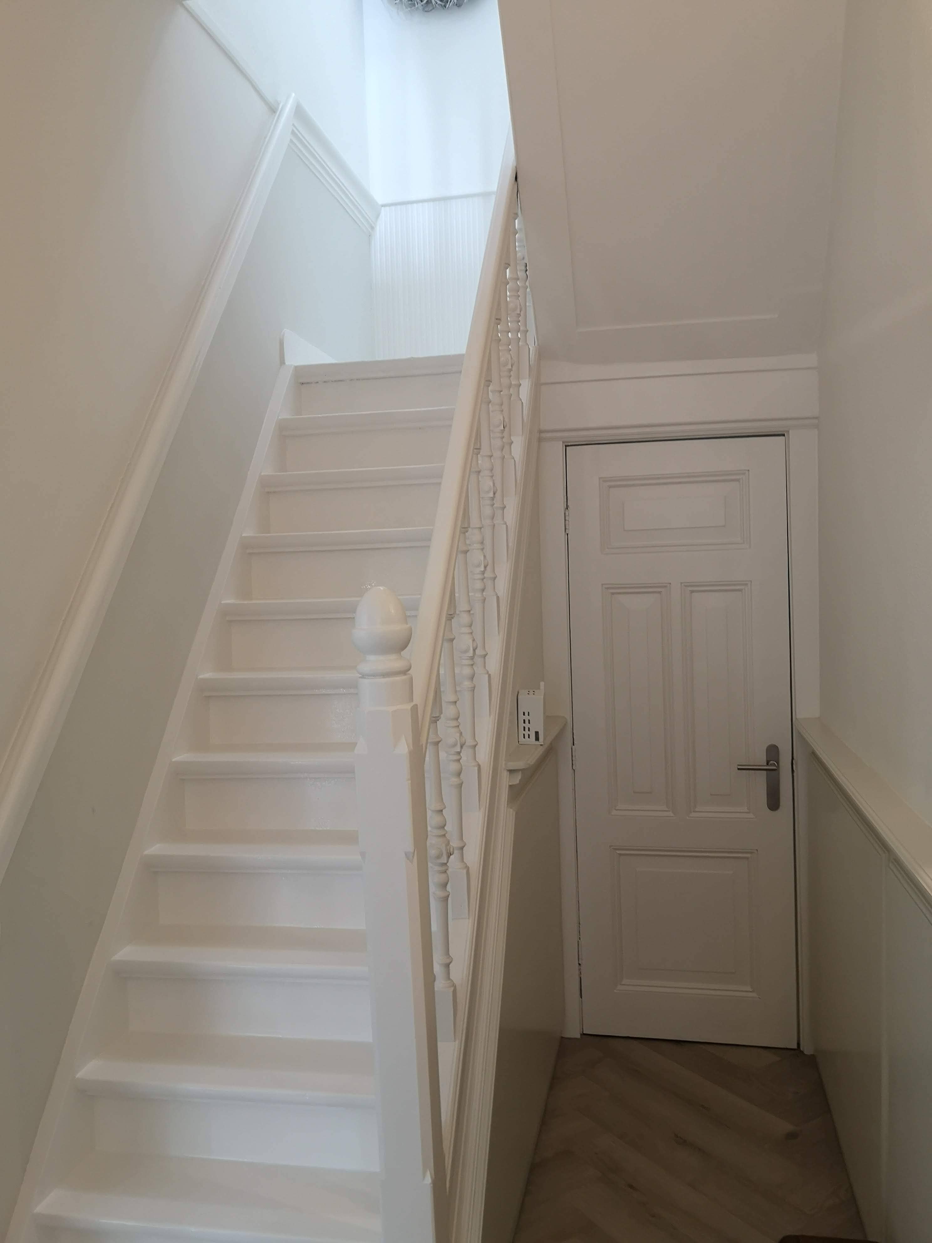 Strak wit geschilderde trap met houten balustrade en overloop in een lichte hal.