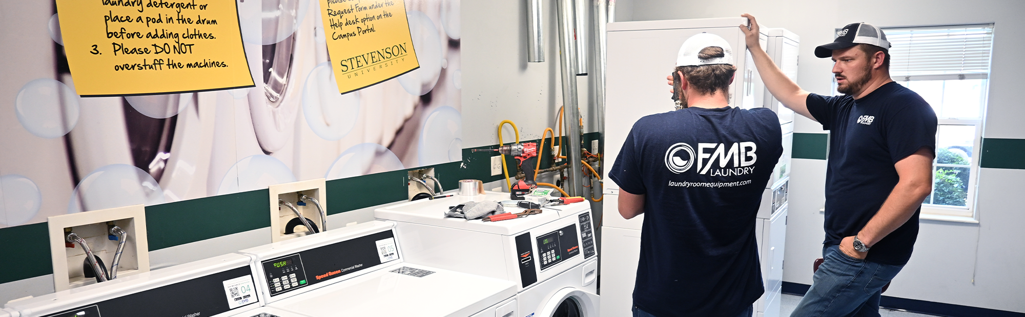 Two FMB Laundry technicians installing commercial washers and dryers in a university residence hall laundry room