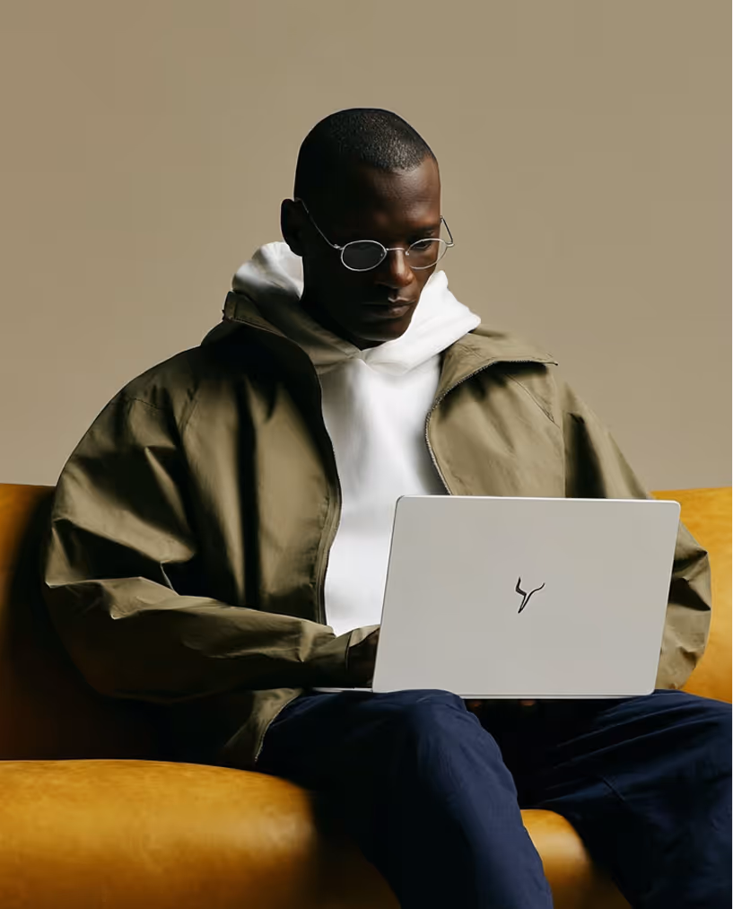 Man wearing glasses and a green jacket over a white hoodie sitting on a yellow couch using a laptop.