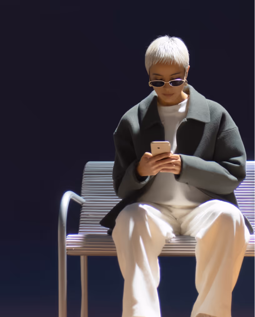 Person with short blonde hair wearing sunglasses and a dark coat sitting on a silver bench while looking at a smartphone.