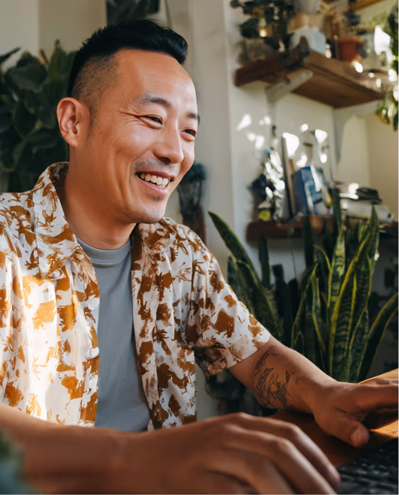 Smiling man with tattoos wearing a patterned shirt typing on a keyboard in a room with plants and shelves.