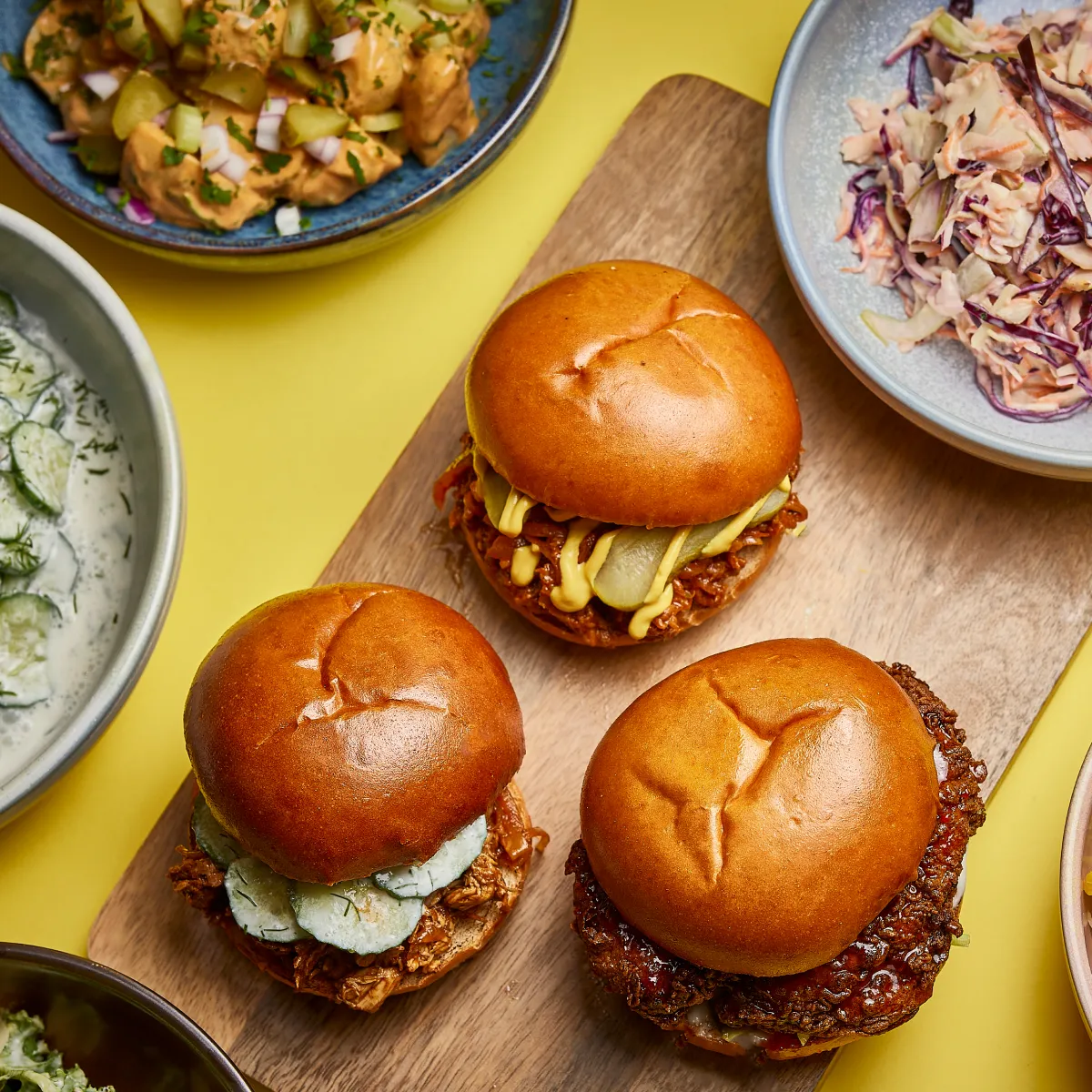 3 burgers on a chopping board with 3 plates of sides.